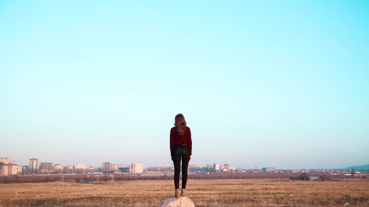 A woman stands alone in a vast open field, gazing towards a distant city skyline under a clear blue sky.